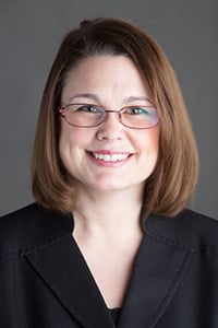 Headshot of Sara Gelser, caucasian brunette woman in a suit and wearing glasses.