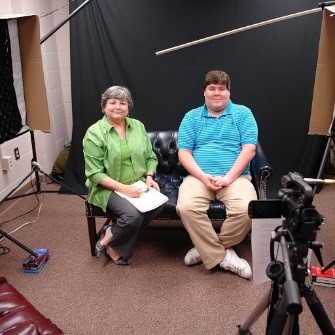 Image of a woman and a man sitting in front of a video camera in front of a backdrop