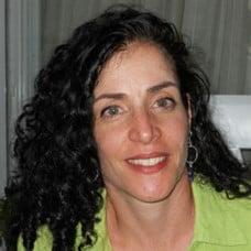 Maya Sabatello, a woman with long curly black hair, she is wearing a collered shirt and smiling at the camera
