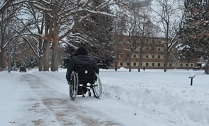 Viewed from the back, a person in a black winter coat rolls his wheelchair down a snowy and tree-lined pathway on what looks to be a college campus.