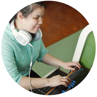 A young Asian woman with hair pulled back wearing a headset around her shoulders and using a braille keyboard. 