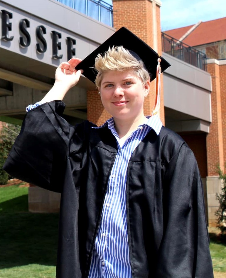 Aley, young white woman with short blonde hair, smiling wearing cap and gown in front of university of Tennessee bridge
