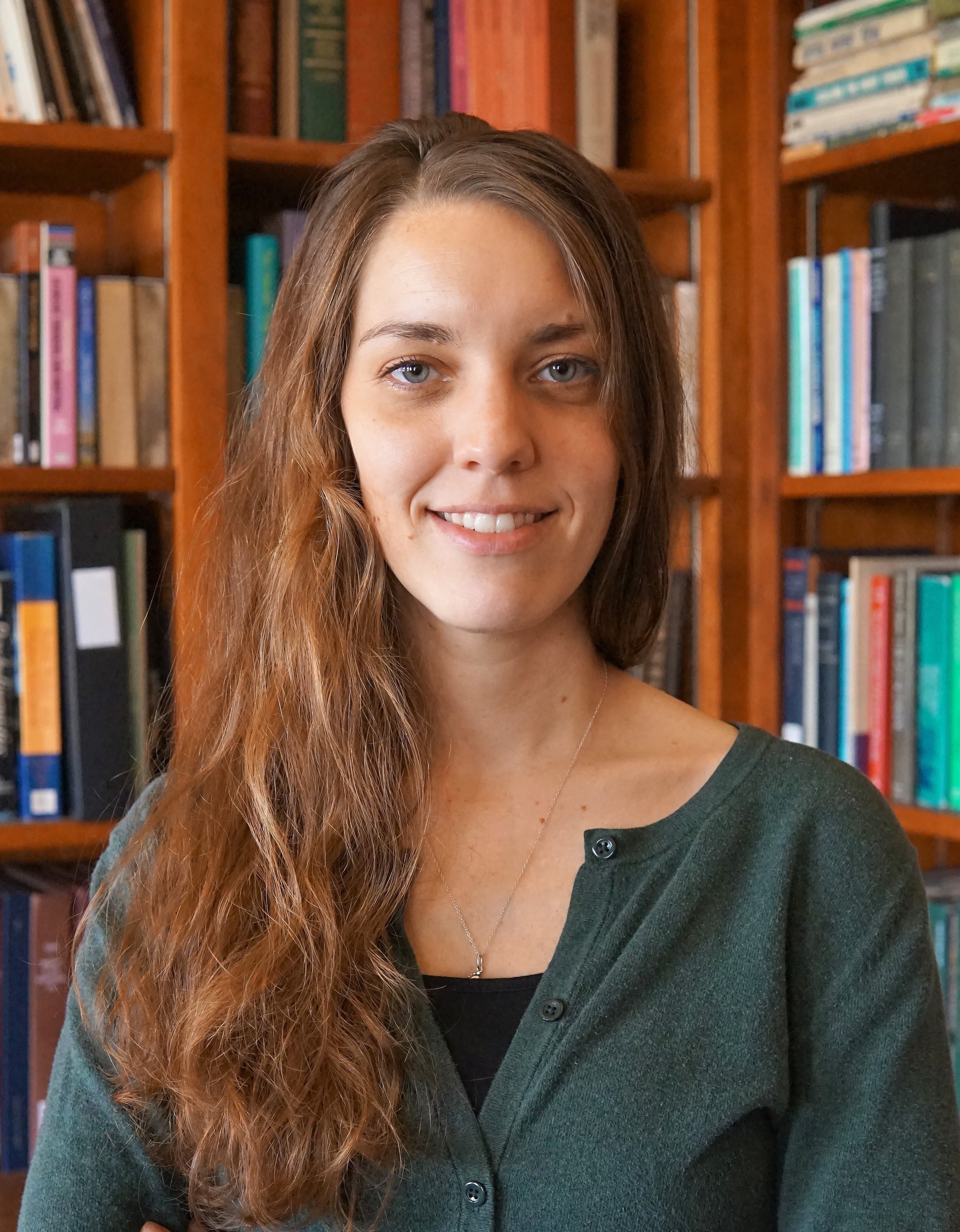 Headshot of Kashia Rosenau, smiling in front of a bookshelf, wearing a dark green pullover shirt with a black tank underneath.  Description automatically generated