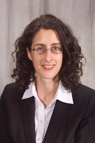 Woman with curly brown hair. She is wearing glasses and a white collared shirt with a black blazer