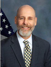 Image of Phil, a white man with a full beard waring a grey suit with checkered yellow tie in front of a blank background and American flag.