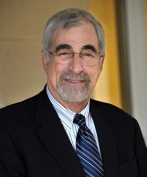 Dr. Riley in a black suit with striped shirt and tie smiling for a headshot with glasses, dark eyebrows, and grey hair and mustache
