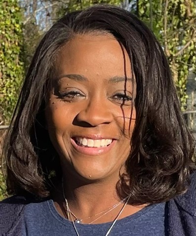 Photo of Donna Johnson, Black woman with long black hair wearing a dark blue blouse around by green plants