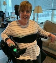 Ellie, a Caucasian woman with short dark brown hair is smiling at the camera. She is wearing a striped shirt and using a standing wheelchair