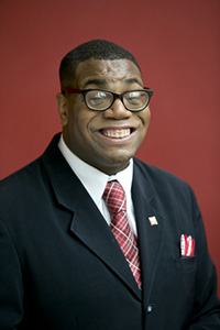 Headshot of Shawn Aelong, black male in a suit and tie, smiling and wearing glasses
