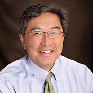 A picture of an Asian man with black and gray hair, parted to the side, sitting, wearing glasses, and smiling at the camera. The image is cutoff at his chest. He is wearing light blue shirt and a green tie with strips. Behind him is a dark background.