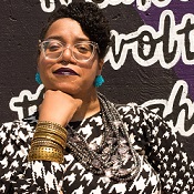 Picture of a honey skinned person sitting in a hot pink and black wheelchair with their hair braided into a swoop across their forehead. They are wearing a black and white houndstooth long sleeve shirt with fire hot pink pants, large clear glasses, Aqua colored earrings that look like rock candy, and a silver multistrand necklace. Their right arm is covered in gold bangles. They are looking fiercely into the camera with their head resting on their right fist. In the background is a purple mural with the word freedom written. 