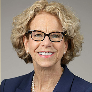 Image of a white woman with short wavy blonde hair wearing glasses and a suit  smiling at the camera.