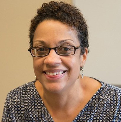 Black woman with short curly brown hair with glasses wearing a blouse and smiling at the camera. 