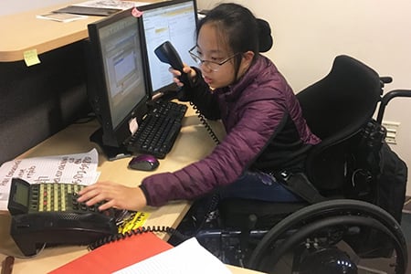Image of an asian women with hairpulled back holdingthe phone reciever and making a call in a office.