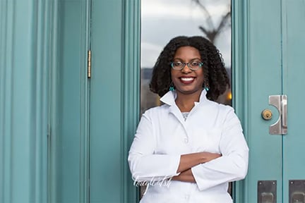 Image of a black woman with shoulder length hair wearing glasses and a white coat smiling at thecamera.
