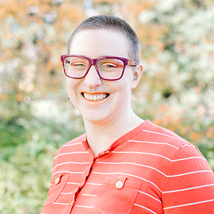 Image of a woman with buzzed hair and wearing bright red glasses with a striped shirt smiling at the camera.and 