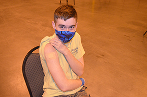 Image of a boy with short brown hair wearing a mask lifting his shortsleeve shirt arm to show a bandaidf from being vaccinated