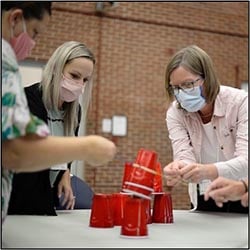 Image of three women wearing face masks standing around a table engaging in activity with solo cups.