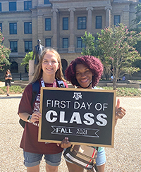 Image of two students standing in front of a college campus holding a sign that says First Day of Class Fall 2021