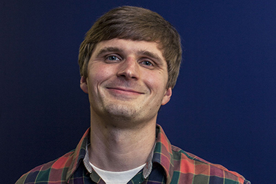 Image of a young white man wearing a flannel shirt smelting at the camera.