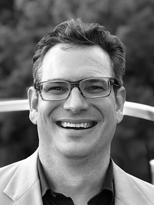 Black and white image of a white man with short brown hair and glasses wearing a shirt and blazer smiling at the camera.