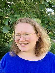  A headshot of white woman wearing a plain blue shirt and gold wire framed glasses. She has shoulder length curly reddish light brown hair and is outside. Leaves and tree branches make up the background.