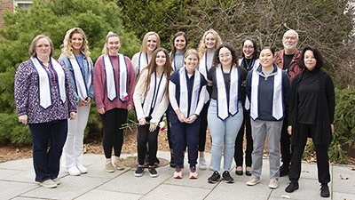 Thirteen people standing outside on a patio for a group photo. Two of the people are college professors; the remaining eleven are college students wearing blue and white graduation stoles that signify their completion of a Disability Studies Minor.  