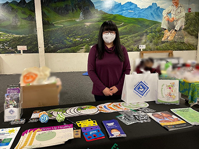 MNLEND Fellow Julie Li Yang is wearing a facemask and standing next to a table with Help Me Grow children's books, toys, and paper resources.