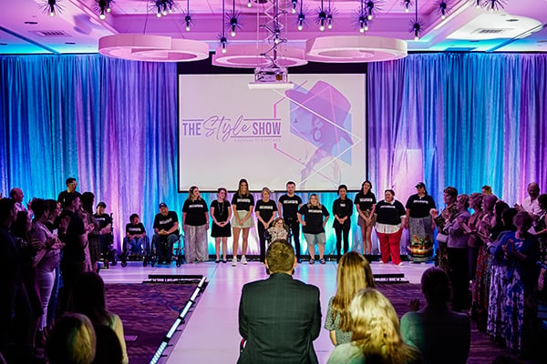 Image of 14 models lined up on the back of a white runway. Behind models are ceiling to floor length curtains with uplighting of pink, purple, blue colors and a project screen with text that says 'The Style Show A Runway to Empower'. Kendra is sitting in front of the line of models. Around the lit up runway are audience members standing and clapping. 