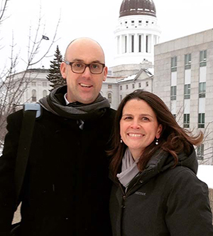 Image of a white man with cropped hair wearing a jacket and scare arm in arm with a white woman with shoulder length haring wearing a scarf and coat standing in front of the Maine State Capitol. 