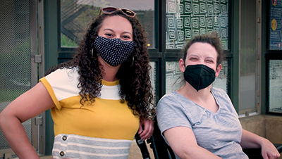A woman with dark curly hair stands next to a woman with hair pulled back, seated in a wheelchair. They are outside and looking at the camera. They are wearing face masks and appear to be smiling