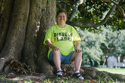 Image of a person wearing a tshirt and shorts sitting on a tree stump and smiling at the camera.