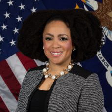 Image of an African American woman wearing a suit and peral knecklaces with the American flage behind her.