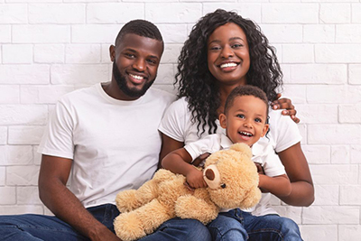 Image of black man and woman holding their child who is also holding at teddy bear everyone is wearing white tshirt and jeans and smiling at the camera.