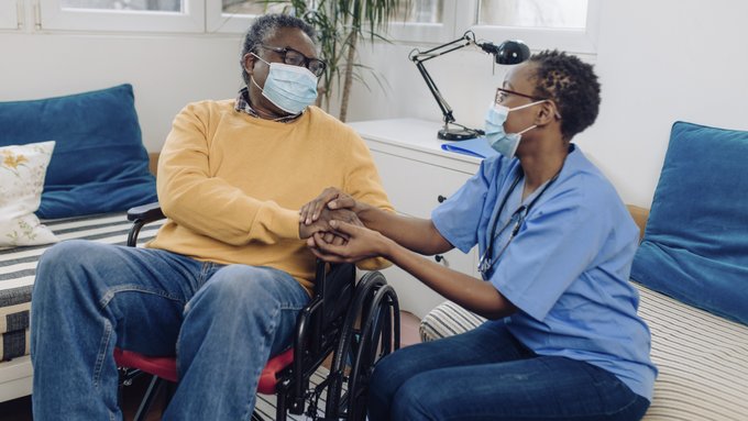Healthcare provider wearing a mask, holding hands with a person another person wearing a mask who is sitting in a wheelchair.
