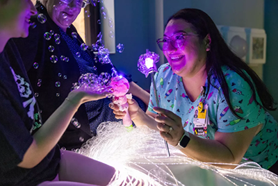 Medical provider making bubles and holding lighting wand with a patient
playing with bubbles and caregiver looking on at the vaccine clini