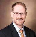 A headshot of Dr. Jeffrey Neul, a smiling white man with short red hair, a beard, and glasses, wearing a black suit jacket, white button-down shirt, and peach tie.