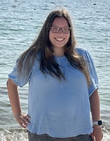 Averie Lane, a woman with long brown hair and glasses wearing a blue blouse standing in front of the ocean.