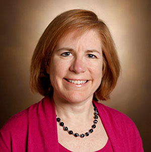 Headshot of Beth Malow, a white woman with chin-length red hair and blue eyes, in front of a brown background. Dr. Malow is wearing a magenta cardigan and camisole set and a necklace
of black beads.
