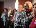 Arie Nettles stands in focus among a line of people. Dr. Nettles is an older Black woman with short platinum hair, black and white cat-eye glasses, pearl earrings, and a black-and-white floral jacket over a black turtleneck.