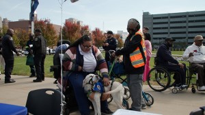 Jamie Junior, an advocacy and community education coordinator for the Disability Network of Wayne County, reaches down to pet a service dog. Jamie is wheelchair bound and is being watched by the dog's owner. The dog's owner is standing to the right o