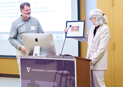  Elise McMillan (gray haired woman wearing a white suit) hands a framed painting to Erik Carter (a tall man with salt and pepper hair and beard wearing a plaid button-down and khaki pants.)