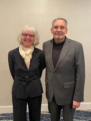 A woman with shoulder-length gray hair is wearing glasses, a pin stripe gray suit, and cream scarf; a man with gray hair and a beard is wearing glasses, a black sweater, and a gray suit.