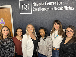 Six female leaders from the Nevada PBISTA stand and smile in front of a blue wall with a white decal of the Nevada Center for Excellence in Disabilities logo. Director, Dr. Ashley Greenwald, is on the far right, and State Coordinator, Kaci Fleetwood, is on the far left.