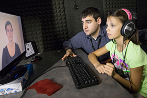 A young male researcher sits next to a young female research participant in front of a computer in a sound booth. The girl is wearing headphones. On the computer screen is a video of a young woman speaking directly to the research participant.
