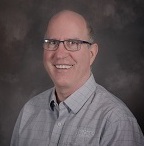 White man smiling at the camera against a gray background. He wears a gray button down shirt. 
