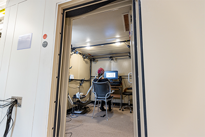 Alt text: Research participant sits in EEG booth, wearing red and black cap while facing a computer screen.