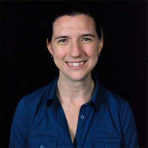 Sarah DeMaio, a woman with brown hair pulled up and wearing a blue shirt, smiles with a black background.