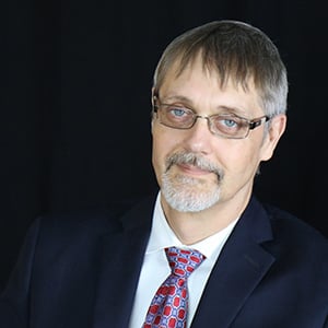 John Tschida, MPP, wearing a white dress shirt, burgundy and gray tie, black suit jacket, gray blonde hair, goatee and glasses.