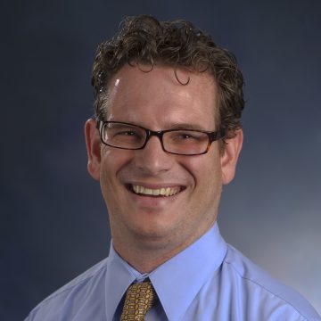 Man with brown curly hair and rectangular glasses smiling a gray background. He wears a blue shirt with a yellow tie. 